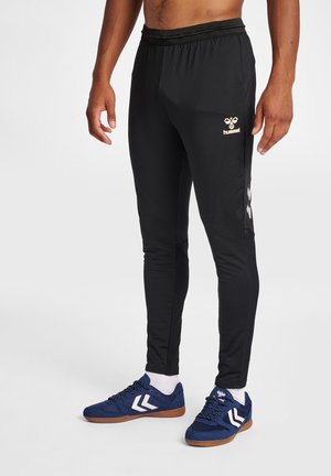 Hombre con pantalones de chándal ajustados negros de hummel y zapatillas azules con logo chevron blanco, de pie frente a un fondo liso.