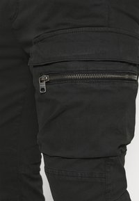 Černé cargo kalhoty s bočním kapsou na zip, vyrobené z hladkého materiálu s pevným designem a prošívanými detaily.