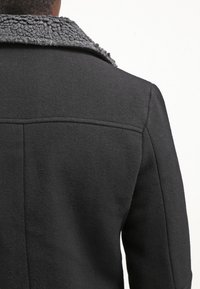 Svart kappa med en texturerad grå krage. Materialet verkar mjukt, med en skräddarsydd passform och synliga sömmar på baksidan.