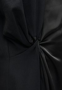 Μαύρο ύφασμα με ματ και σατέν υφές, με μια στριφτή λεπτομέρεια στο κέντρο, δημιουργώντας οπτική αντίθεση και διάσταση.