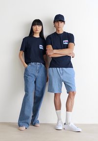 Kvinde i løse jeans og navy t-shirt med applikation står ved siden af en mand i lyse denimshorts, navy t-shirt, kasket, hvide strømper og sneakers.