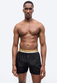 Αθλητικός άνδρας όρθιος, φοράει μαύρα μπόξερ Calvin Klein με κίτρινη μέση, με ουδέτερη έκφραση, σε απλό ανοιχτό φόντο.