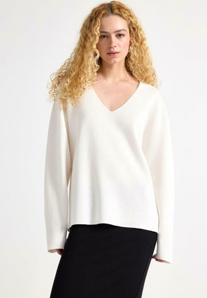Pull blanc à col en V avec des manches longues, coupe décontractée, texture douce. Porté avec une jupe noire, à coupe droite et au design minimaliste.