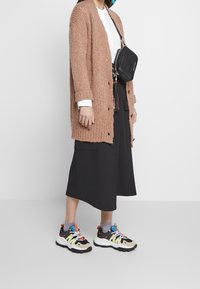 Hellbraun gestrickte Strickjacke, weißes Hemd, schwarze weit geschnittene Hose und mehrfarbige Sportschuhe mit klobigen Sohlen. Schwarze Umhängetasche.