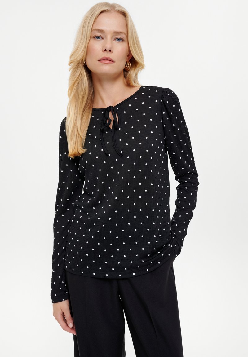 Czarna bluzka z długim rękawem w białe kropki, okrągły dekolt oraz zawiązywany detal przy szyi. Miękki, strukturalny materiał z marszczonymi rękawami.