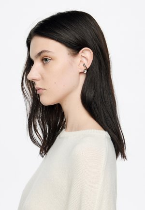 Pendientes de plata con un diseño curvado que presenta un acento esférico claro, llevados por una modelo con el cabello oscuro y liso, vestida con un suéter ligero.