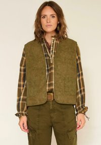 Femme portant un gilet en cuir suédé vert olive, une chemise à carreaux avec des poignets volantés, un pantalon cargo vert olive, se tenant devant un fond blanc cassé.