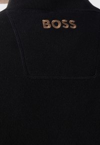 Gros plan sur un vêtement en maille noire avec texte brodé en majuscules marron « BOSS » près du haut du milieu du dos sous le col.