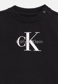 T-shirt noir en coton avec un grand logo "CK" blanc et un texte "Calvin Klein" plus petit. Comprend deux boutons pressions à l'épaule pour faciliter l'enfilage.