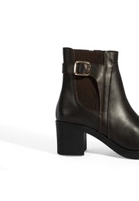 Bruine leren enkelboot met een groef elastische paneel, een metalen gesp accent en een stevige zwarte hak. Gladde textuur met minimale stiksels.