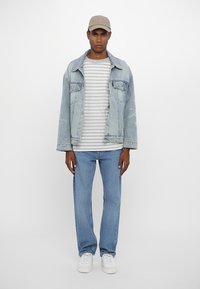 Light blue denim jacket over a gray and white striped shirt, paired with blue jeans and white sneakers. The model wears a beige cap.