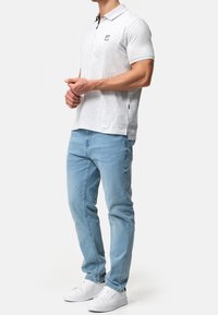 Polo shirt grigio chiaro con colletto e piccolo logo, abbinato a jeans azzurri e sneakers bianche, con spacchi laterali e una vestibilità casual.