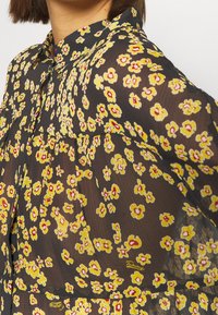 Blusa nera a fiori con piccoli fiori gialli e centri rossi, colletto e tessuto trasparente con una sottile trama.