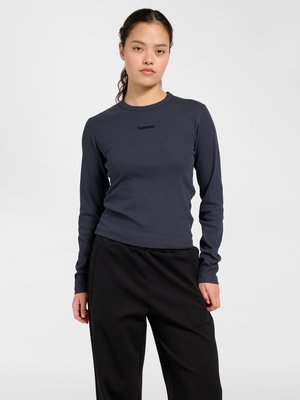 Młoda kobieta z ciemnymi włosami spiętymi do tyłu, ubrana w dopasowaną ciemnoszarą bluzkę z długim rękawem i luźne czarne spodnie, stoi na białym tle.