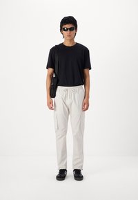 Schwarzes T-Shirt, hellbeige Cargo-Hosen mit Seitentaschen, schwarze Umhängetasche und schwarze Sneakers; Person trägt Sonnenbrille und steht vor einem einfarbigen Hintergrund.