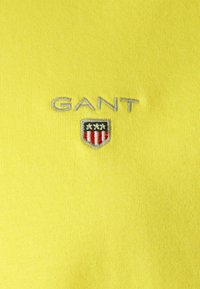 Geel katoenen poloshirt met geborduurd "GANT" logo in zilver en een schildpatch met rode en zwarte accenten. Glad textuur.