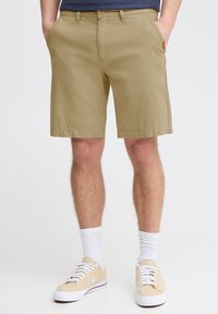 Beige katoenen short met een rechte voorkant, twee zijzakken en lussen voor een riem. Draagt met lichte sneakers en witte sokken.