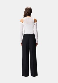 Donna con lunghi capelli castani che indossa un top bianco a maniche lunghe scollato sulle spalle e pantaloni neri a gamba larga e vita alta, vista da dietro.