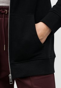 Black zip-up sweatshirt with ribbed cuffs and pockets, paired with burgundy pants featuring a textured fabric and drawstring.