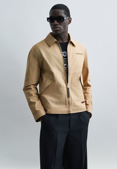 Homme portant des lunettes de soleil noires, une veste zippée beige sur un t-shirt noir à motif, et un pantalon sombre plissé, mains dans les poches, regardant vers la gauche.
