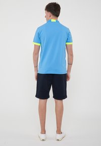 Polo blu con colletto giallo e accenti sulle maniche, abbinato a pantaloni corti navy arrotolati e scarpe bianche. Tessuto morbido, design casual.