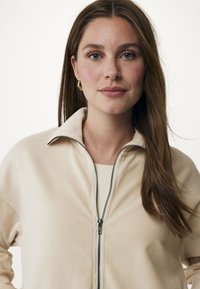 Ljusbeige zip-jacka med ståkrage, med en texturerad tyg och svarta dragkedjedetaljer. Enkelt design med en avslappnad passform.