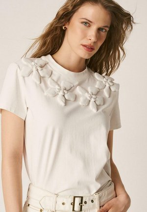 Mujer con camiseta blanca de manga corta con aplicaciones florales 3D en los hombros, combinada con pantalones blancos de talle alto y cinturón.