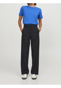 Blå kortärmad t-shirt och svarta högmidjade byxor med rak passform. Vita sneakers med mörka sulor, händerna i fickorna.