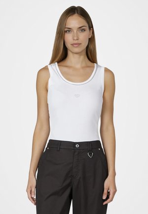 Woman with long straight hair wearing a white sleeveless top and black pants, standing against a plain light background.