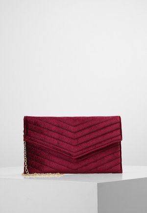 Bolso sobre de terciopelo rojo oscuro con patrón de costura en chevron y correa de cadena dorada, reposando sobre un pedestal blanco.