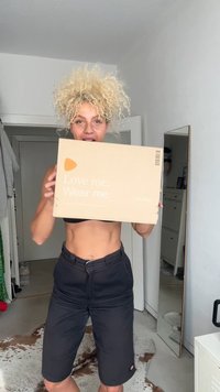Caixa de cartão castanho com o texto "Ame-me. Vista-me." e um triângulo laranja. A caixa é segurada por uma pessoa vestindo uma parte de cima preta e calções.