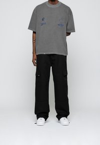 Person mit einem grauen, oversize Grafik-T-Shirt, schwarzen Cargo-Hosen mit Taschen und weißen Turnschuhen, der vor einem schlichten weißen Hintergrund steht.