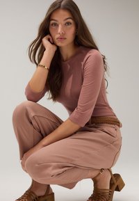 Soonikkoes, pikkade varrukatega tolmuroosa pluus laia lõikega heledate roosade pükstega. Märkimisväärsed detailid hõlmavad punutud pruuni vööd ja pruune kontsakingi.