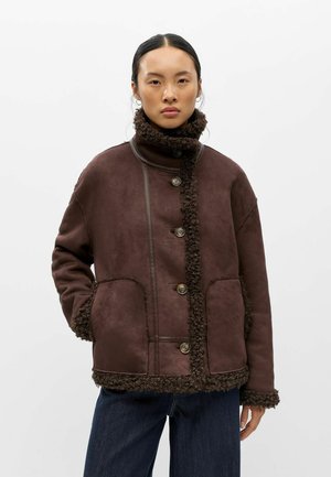 Jeune femme portant une veste en shearling marron avec de grands boutons, deux poches avant et un jean bleu foncé sur un fond uni.