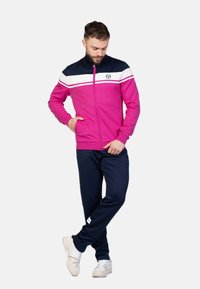 Trainingsanzug mit einer rosa Reißverschlussjacke, die weiße und marineblaue Streifen hat, kombiniert mit marineblauen Hosen. Hergestellt aus leichtem, dehnbarem Material.