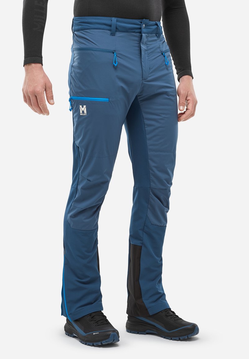 Homme portant un pantalon de plein air bleu avec des poches zippées, des chaussures de randonnée noires et un haut à manches longues noir, se tenant sur un fond blanc.
