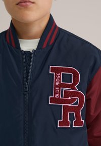 Ung person klädd i en marinblå och vinröd varsityjacka med dragkedja och texturerad "BR"-patch broderad med "Blue Ridge" på bröstet.