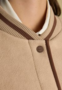 Beež fliiskinnas, millel on ribitud krae ja pruuni ja kreemikaid triibusid. Silmapaistev pruun nupp logodetailiga kaela juures.