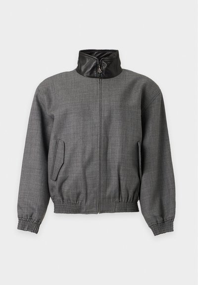 Veste grise zippée avec col montant en cuir noir, poignets élastiques et deux poches latérales à rabat sur fond uni.