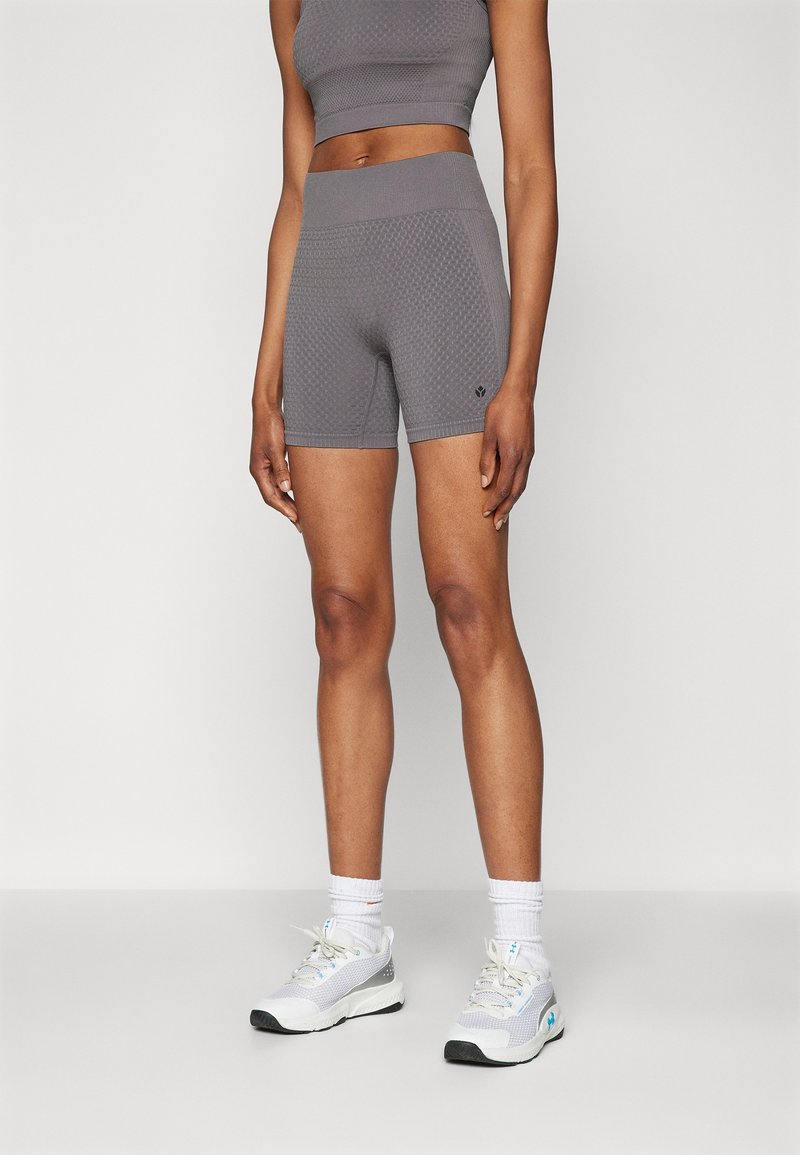 Femme portant un short cycliste gris texturé taille haute et un top court assorti, associés à des chaussures de sport blanches et des chaussettes, debout sur un fond uni.