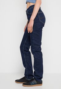 Personne portant un jean droit bleu foncé, des baskets noires avec des rayures bleues, et un haut court bleu clair, debout de profil.