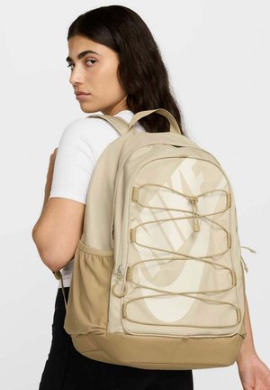 Mochila Nike beige con un gran logo, tela texturizada, bolsillo de malla lateral, cierres de cremallera y correas ajustables.