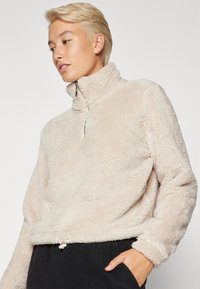 Pull en polaire beige avec un col montant et une fermeture à demi-zip, présentant une surface douce et texturée ainsi qu'un cordon de serrage à la taille ajustable.