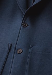 Dunkelblaue Jacke aus strukturiertem Stoff, mit zwei Taschen und zwei schwarzen Knöpfen an der Vorderseite, sowie einem glatten Kragendesign.