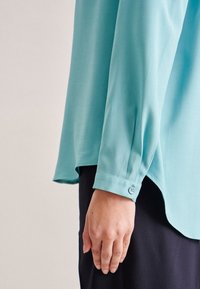 Camicia azzurro chiaro a maniche lunghe con polsini abbottonati, realizzata in tessuto liscio. L'orlo è leggermente curvo, abbinata a pantaloni scuri.