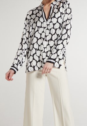 Donna che indossa una camicetta a maniche lunghe con motivo bianco e nero abbinata a pantaloni bianchi a vita alta e gamba larga, su sfondo semplice.