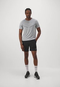 Sportos férfi áll, egyik kezével a zsebében, szürke rövid ujjú pólót, fekete sortot, fehér Nike zoknit és fekete futócipőt visel.