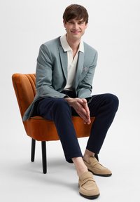 Lichtblauwe blazer over een witte polo, marineblauwe broek en beige instappers. De stoel is van oranje fluweel met zwarte poten; de achtergrond is vlak wit.