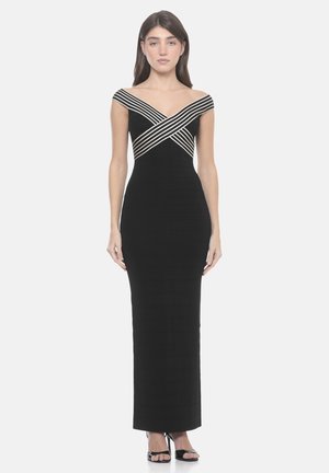 Donna in piedi che indossa un lungo abito nero aderente con spalline a righe scoperte che si incrociano sul petto, abbinato a tacchi neri aperti in punta.