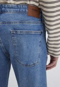 Blue denim jeans with a leather brand patch reading "mavi." Features include visible stitching, back pockets, and a relaxed fit. Striped shirt sleeve visible.